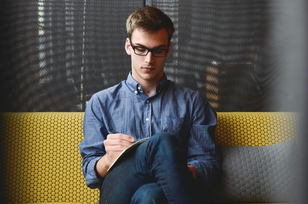 entrepreneur-startup-start-up-man-39866-39866 A young man in glasses writes in a notebook while sitting on a stylish couch indoors.